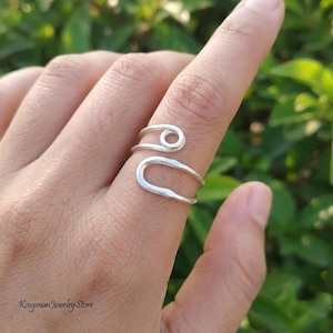 May include: A silver-colored ring featuring an open, bypass-style design. The band consists of two distinct, smooth wire-like segments that wrap around the finger, with one end forming a small circular loop and the other a larger, elongated U-shape.