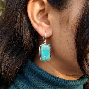 May include: A rectangular turquoise gemstone earring set in a silver frame with a twisted rope border. The stone features natural blue and green hues with brown matrix veining. The earring hangs from a simple silver hook wire attachment.