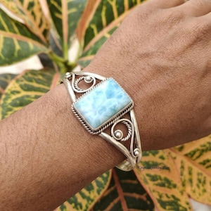 May include: A silver cuff bracelet featuring a rectangular, light blue Larimar stone set in a rope-textured frame. The band is crafted from polished silver wire with decorative swirling scrollwork patterns on both sides of the central stone.