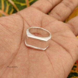 Könnte beinhalten: Ein polierter Silberring mit einer breiten, flachen, rechteckigen Vorderseite und einer glatten, massiven Ringschiene. Die Metalloberfläche ist reflektierend und schlicht und zeigt ein klares, geometrisches Design mit scharfen Kanten und einem gleichmäßigen Finish.