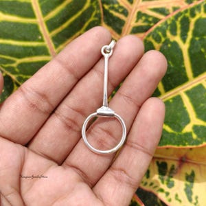 Puede incluir: Un colgante de metal plateado con forma de bocado de caballo, que presenta una barra vertical conectada a un anillo circular en la parte inferior. La pieza tiene un acabado liso y pulido, e incluye una pequeña anilla en la parte superior.