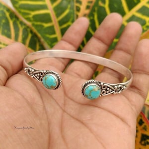 May include: A silver cuff bracelet featuring two round, bright blue turquoise stones with brown matrix patterns. The metal band displays intricate, decorative scrollwork and twisted rope detailing surrounding each stone setting at the open ends.