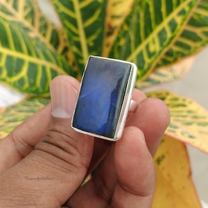 May include: A rectangular labradorite gemstone ring set in a polished silver-toned metal band. The stone features a deep blue iridescent flash across its smooth, flat surface. The ring is held between fingers, showcasing the stone's reflective quality.