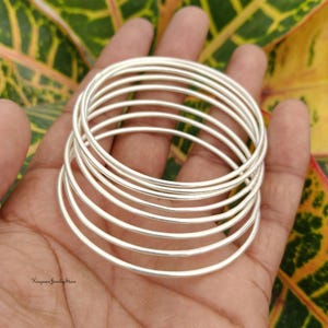 Puede incluir: Un conjunto de siete brazaletes finos de color plateado pulido apilados juntos. Las pulseras circulares presentan una superficie metálica lisa y reflectante con un perfil redondeado simple. El texto Kingman Jewelry Store es visible en la esquina.