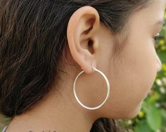 May include: A simple, polished silver hoop earring with a circular design and a thin wire post closure. The earring is worn in the earlobe, featuring a smooth, reflective metallic surface that catches the light, creating a clean and classic appearance.