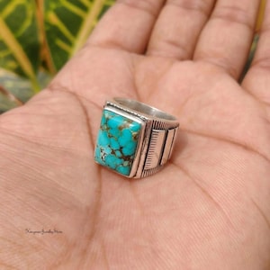 May include: A silver ring featuring a rectangular turquoise stone with a brown matrix pattern. The band has a polished finish with engraved vertical line details on the sides. The text Kinyman Jewelry Store is visible in the bottom left corner.