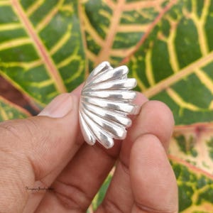 Puede incluir: Un anillo de plata pulida con un diseño en forma de abanico que presenta múltiples crestas redondeadas y elevadas que irradian desde la base. La superficie metálica es lisa y reflectante, mostrando un patrón distintivo de concha.