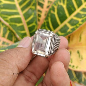 Pode incluir: Um anel de prata com uma grande pedra preciosa retangular transparente e facetada, montada numa banda de metal texturizado. A banda apresenta um padrão detalhado de hachuras cruzadas nas laterais. O texto Kingman Jewelry Store é visível.