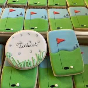 May include: A collection of decorated sugar cookies featuring a golf course theme. The cookies are decorated with green icing to represent grass, blue icing to represent the sky, and white icing to represent golf balls. One cookie features a golf ball with the word "Titleist" written on it.