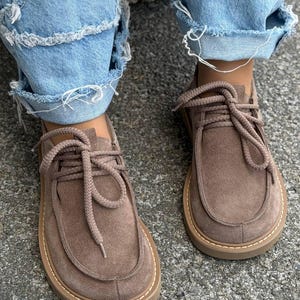 Könnte beinhalten: Ein Paar taupefarbene Wildlederschuhe mit abgerundeter Spitze, dicken seilartigen Schnürsenkeln und einer flachen Gummisohle mit sichtbaren hellen Nähten am Rand. Die Schuhe haben ein niedriges Design mit einer mokassinartigen Konstruktion.