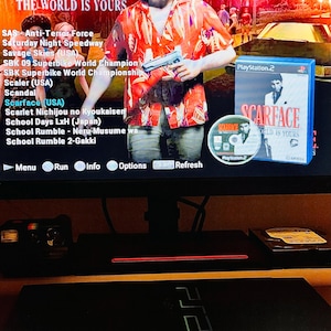 May include: A black PlayStation 2 console sits below a monitor displaying the Scarface: The World Is Yours video game menu. The screen shows a list of game titles, a character in a red patterned shirt holding a gun, and the game case and disc.