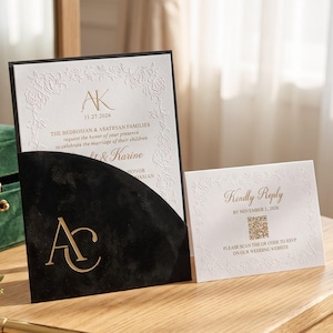 May include: A white wedding invitation card with gold text and embossed floral borders, tucked into a black velvet pocket featuring a gold monogram. Beside it, a matching white RSVP card with gold text and a QR code for wedding website access.