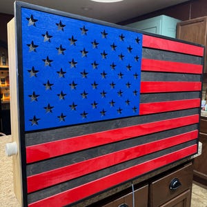Peut inclure: Décor mural en bois représentant le drapeau américain. Il présente un champ bleu avec des étoiles découpées, des rayures rouges et noires. Le cadre est noir, et l'ensemble est monté sur une base en bois clair. Un élément de décoration patriotique.