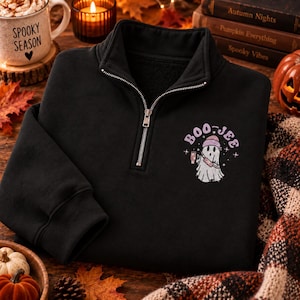 May include: A black quarter-zip sweatshirt featuring a graphic on the left chest of a white ghost wearing a pink beanie and holding a drink, with the text BOO-JEE printed in a curved purple font above the ghost, surrounded by small star accents.