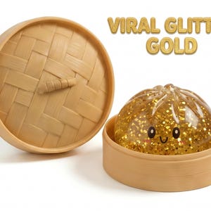 May include: A clear, gold glitter-filled dumpling toy with a smiling face, resting inside a light brown bamboo steamer basket with a matching lid. The text VIRAL GLITTER GOLD is printed in the upper right corner in a gold, bubbly font.