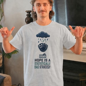 May include: A light gray short-sleeve t-shirt featuring a dark blue graphic of a rain cloud over a baseball glove and a scoreboard. Below the graphic, the text reads: HOPE IS A STATISTICALLY BAD STRATEGY in dark blue and teal capital letters.