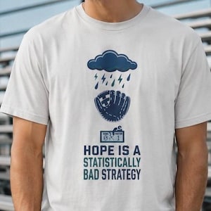 May include: A white short-sleeved t-shirt featuring a dark blue graphic of a rain cloud above a baseball glove and a scoreboard displaying 0-1. Below the graphic, the text reads: HOPE IS A STATISTICALLY BAD STRATEGY in dark blue capital letters.