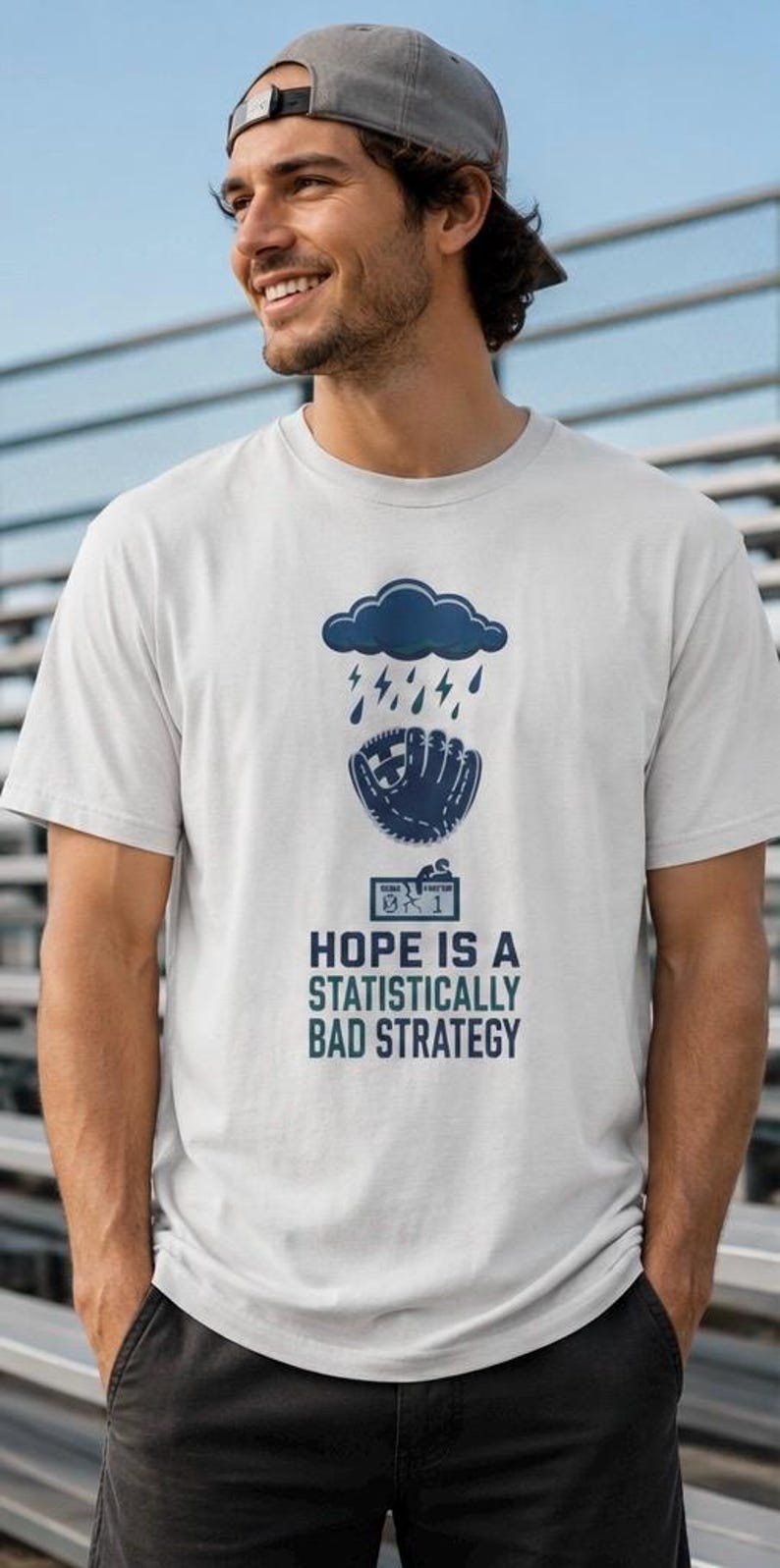 May include: A white short-sleeved t-shirt featuring a dark blue graphic of a rain cloud above a baseball glove and a scoreboard displaying 0-1. Below the graphic, the text reads: HOPE IS A STATISTICALLY BAD STRATEGY in dark blue capital letters.