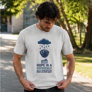 May include: A light gray short-sleeved t-shirt featuring a dark blue graphic of a rain cloud over a baseball glove and a scoreboard. Below the graphic, the text reads: HOPE IS A STATISTICALLY BAD STRATEGY in dark blue capital letters.