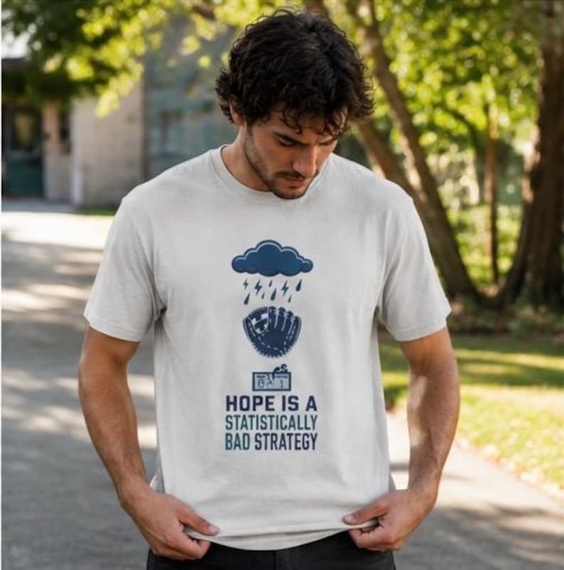May include: A light gray short-sleeved t-shirt featuring a dark blue graphic of a rain cloud over a baseball glove and a scoreboard. Below the graphic, the text reads: HOPE IS A STATISTICALLY BAD STRATEGY in dark blue capital letters.