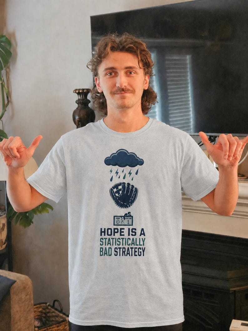 May include: A light gray short-sleeve t-shirt featuring a dark blue graphic of a rain cloud over a baseball glove and a scoreboard. Below the graphic, the text reads: HOPE IS A STATISTICALLY BAD STRATEGY in dark blue and teal capital letters.