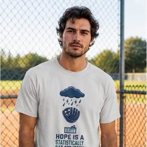 May include: A light gray short-sleeve t-shirt featuring a dark blue graphic of a rain cloud with lightning bolts above a baseball glove. Below the glove is a scoreboard showing 0-1 and the text: HOPE IS A STATISTICALLY BAD STRATEGY.