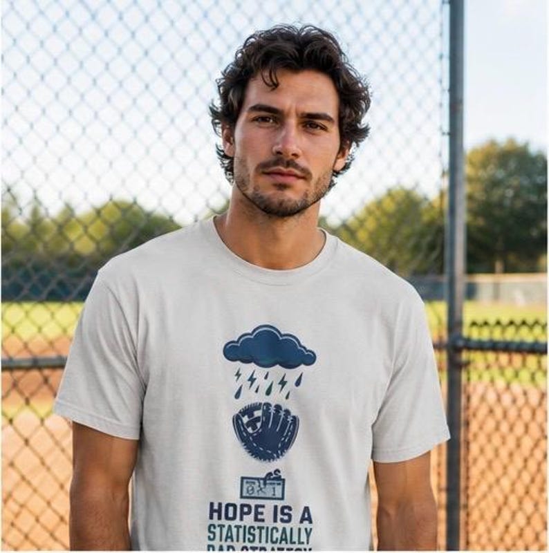 May include: A light gray short-sleeve t-shirt featuring a dark blue graphic of a rain cloud with lightning bolts above a baseball glove. Below the glove is a scoreboard showing 0-1 and the text: HOPE IS A STATISTICALLY BAD STRATEGY.