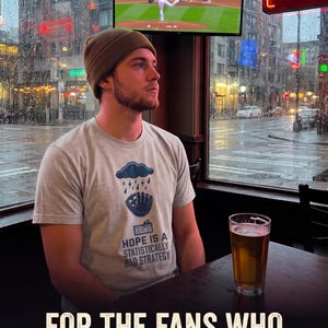 May include: A light gray short-sleeved t-shirt featuring a graphic of a rain cloud over a baseball glove with the text "HOPE IS A STATISTICALLY BAD STRATEGY" printed below. The shirt is worn with a solid brown beanie in a dimly lit indoor setting.