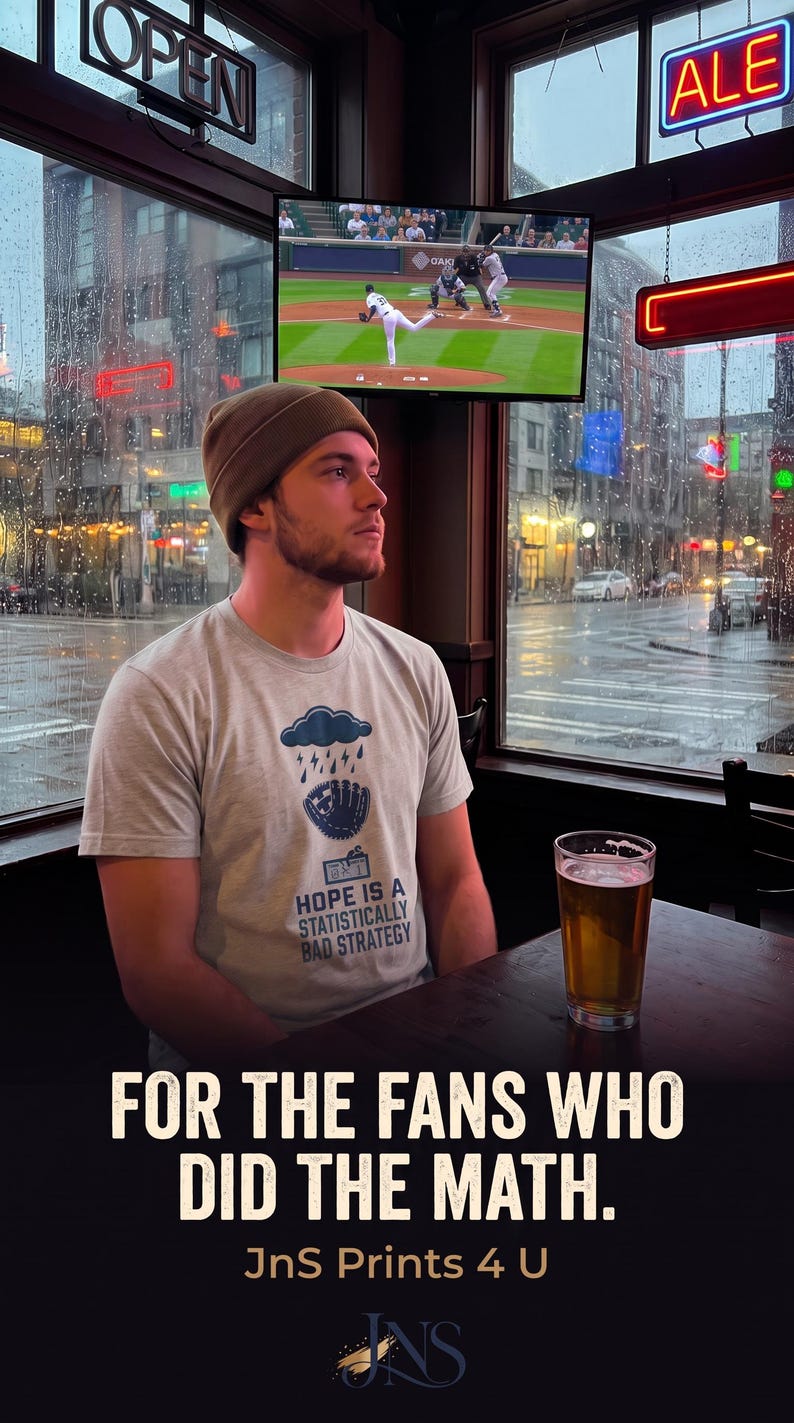 May include: A light gray short-sleeved t-shirt featuring a graphic of a rain cloud over a baseball glove with the text "HOPE IS A STATISTICALLY BAD STRATEGY" printed below. The shirt is worn with a solid brown beanie in a dimly lit indoor setting.
