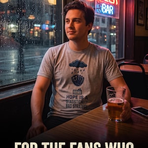 May include: A light gray short-sleeve t-shirt featuring a dark blue graphic of a rain cloud over a baseball glove, with the text "HOPE IS A STATISTICALLY BAD STRATEGY" printed below the graphic in a dark blue sans-serif font.