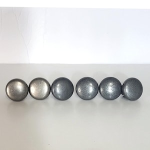 May include: A set of six circular cabinet knobs with a smooth, metallic finish in varying shades of dark gray and silver. Each knob features a rounded, dome-shaped top and a polished surface that reflects light, arranged in a horizontal row.
