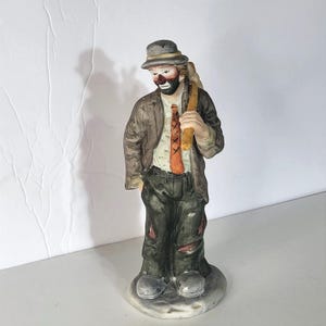 Può includere: Una statuetta in ceramica di un clown vagabondo che indossa un cappello grigio, una giacca marrone, una camicia bianca, una cravatta arancione con cuciture nere e pantaloni verde scuro. Il clown ha il viso dipinto con un naso rosso.