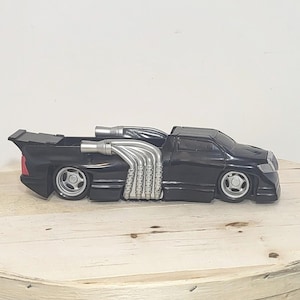 May include: A black toy truck featuring a prominent silver exhaust pipe assembly along the side, a rear spoiler, and silver wheels. The vehicle has a low-profile design with a sleek body, clear headlights, and a red taillight visible at the rear.