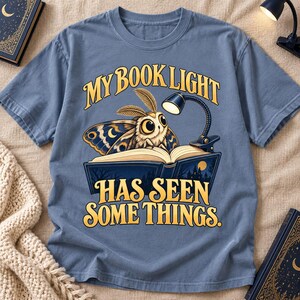 May include: A slate blue t-shirt featuring a graphic of a moth with large eyes reading an open book under a clip-on lamp. The text reads MY BOOK LIGHT HAS SEEN SOME THINGS in yellow lettering. The design includes a dark blue book cover.