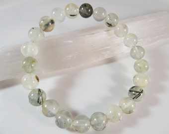 May include: A bracelet made of translucent, round beads with black and green inclusions. The beads are strung together, forming a circular shape. The bracelet is displayed on a white surface with a white crystal in the background.