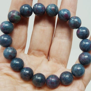 Op de afbeelding: Een armband met ronde, gepolijste kralen in blauwe en rode tinten. De kralen zijn aan elkaar geregen tot een complete cirkel. De armband wordt op een hand getoond.