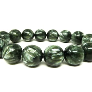 Puede incluir: Un brazalete hecho de cuentas redondas y pulidas de serafinita. Las cuentas son de color verde oscuro con inclusiones plateadas y plumosas, creando un patrón único. El brazalete está ensartado, mostrando la belleza natural de la piedra preciosa.
