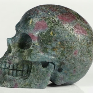 May include: A carved skull made of a stone material, featuring a mix of green and red hues. The skull has detailed facial features, including eye sockets, a nose cavity, and a row of teeth. The stone has a textured surface.