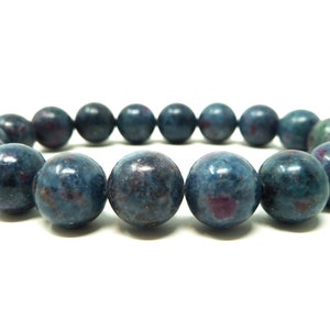 Puede incluir: Una pulsera de cuentas redondas y pulidas. Las cuentas son principalmente de color azul oscuro con motas rojas y moradas. La pulsera se muestra sobre un fondo blanco. Cada cuenta mide aproximadamente 1 cm de diámetro.