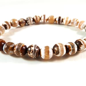 Puede incluir: Pulsera de cuentas redondas en tonos marrón, beige y blanco. Las cuentas tienen un diseño a rayas y con patrones. La pulsera está ensartada en un cordón elástico. Cada cuenta mide aproximadamente 0,8 cm de diámetro.