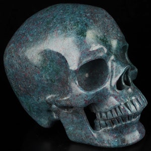 May include: A detailed side view of a stone skull. The skull is a mottled teal color with red and white flecks. The skull's features, including the eye sockets, nasal cavity, and teeth, are clearly defined. The background is solid black.