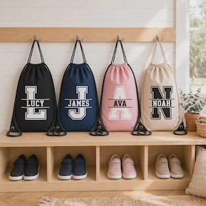 Peut inclure: Quatre sacs à dos à cordon en noir, bleu marine, rose et beige, chacun arborant une grande lettre de style universitaire avec une ligne horizontale et un nom imprimé : LUCY, JAMES, AVA et NOAH. Chaque sac possède des cordons noirs.
