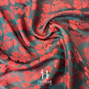May include: A dark teal fabric featuring a repeating pattern of vibrant red flowers. The material has a subtle sheen and folds throughout the surface. At the bottom center, the text "Hà Hoat Silk" is printed in a white, stylized font.