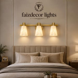 Pode incluir: Uma luminária de parede de três luzes com uma barra de metal dourado escovado e três cúpulas cónicas de vidro branco translúcido. A luminária está montada sobre uma cama e apresenta o texto faizdecor lights com um logótipo acima.