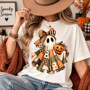 May include: A white t-shirt featuring a graphic of a ghost wearing a large plaid bow. The ghost is depicted with a patchwork pattern in shades of orange, green, and black, holding a small orange jack-o'-lantern with a heart-shaped tag.