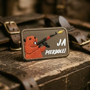 May include: A rectangular olive green embroidered patch featuring an orange beaver holding a black rifle with a yellow muzzle flash. The text JA PIERDOLEI is stitched in white letters on the right side of the patch, which has a dark border.