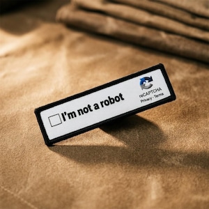 Könnte beinhalten: Ein rechteckiger gestickter Aufnäher mit weißem Hintergrund und schwarzem Rand. Der Aufnäher zeigt ein schwarzes quadratisches Kontrollkästchen, gefolgt vom Text I'm not a robot in schwarzer Schrift sowie dem reCAPTCHA-Logo und Privacy - Terms.