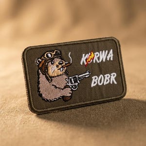 Può includere: Una toppa rettangolare ricamata color verde oliva raffigurante un castoro dei cartoni animati con occhiali da aviatore, che fuma un sigaro e impugna un revolver. Il testo "KURWA" con un fulmine e "BOBR" è ricamato in filo bianco a destra.