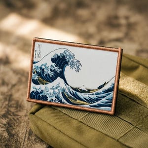 May include: A rectangular morale patch featuring a printed reproduction of The Great Wave off Kanagawa. The design displays a large dark blue wave with white foam crests, a small mountain in the background, and tan border stitching.