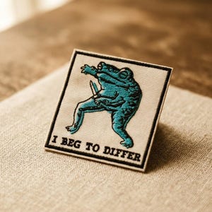 May include: A square embroidered patch on a light beige fabric background. The patch features a teal frog holding a small knife, depicted in a dynamic pose. Below the frog, the text I BEG TO DIFFER is embroidered in bold black capital letters.
